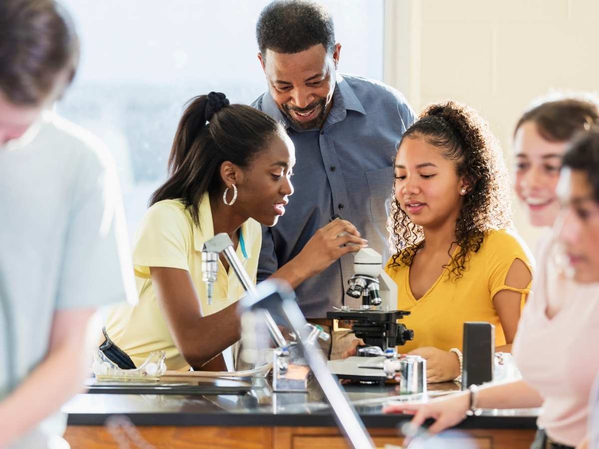 High school kids working in a science lab with a teacher overlooking as they use a microscope for article Channelview ISD