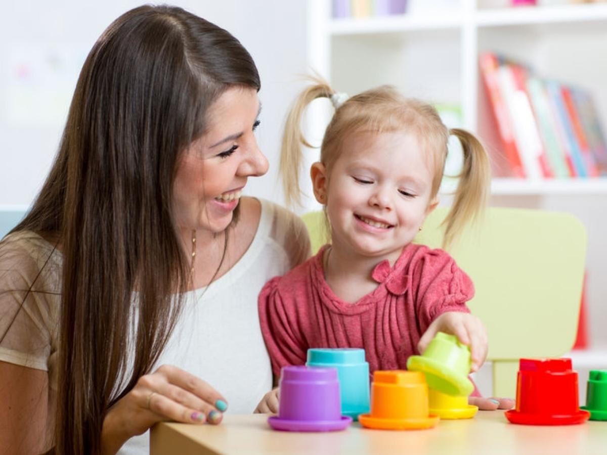 Teacher kneeling down smiling at child who is playing with colored cups for article Primrose Schools - Northeast Houston