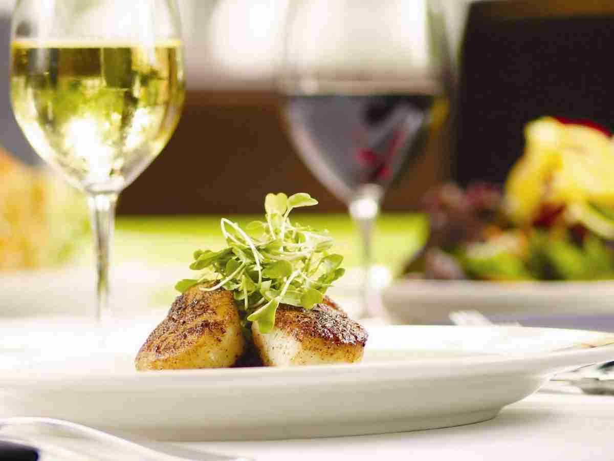 Scallops plated fancy with green leafy item on top and glass of white wine in background from Steakhouse on the Bayou for article Houstons Diverse Dining Scene