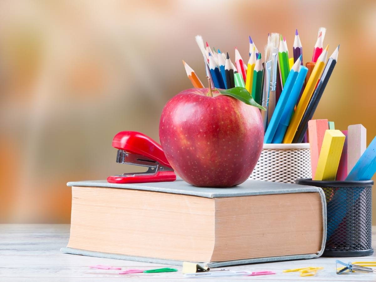 Close up of a book with an apple and stapler on it and a container of colored pencils and one of colored chalk behind it for article Klein Independent School District