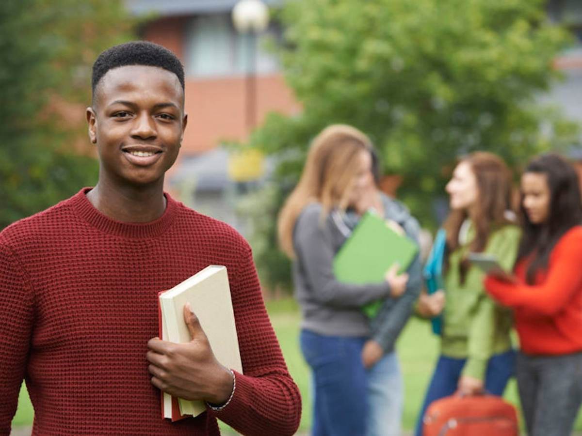 Male college student holding books looking at camera with female students in the background in a group talking to each other for article San Jacinto College