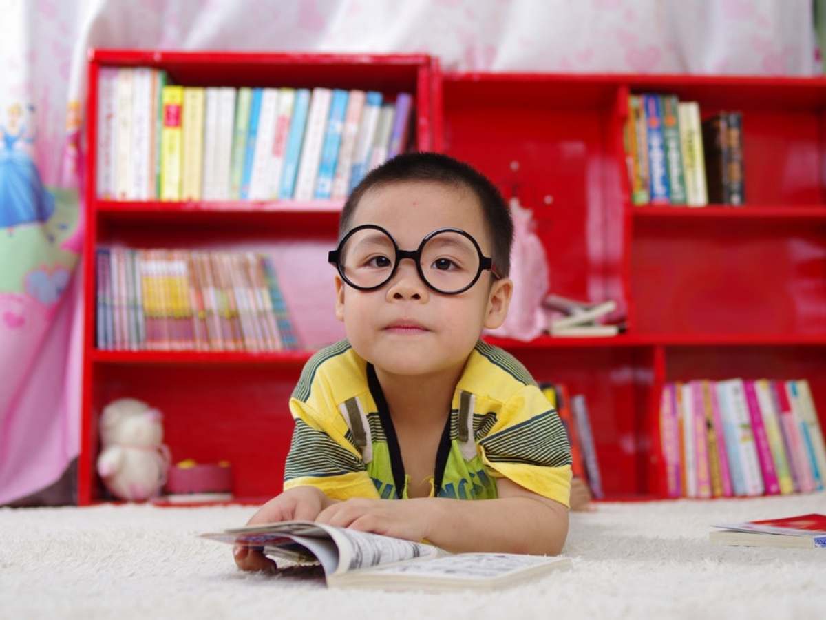 Young child with glasses on laying on carpeted floor reading a book with a bright red bookcase full of books behind him for article Public Schools in Fort Bend County