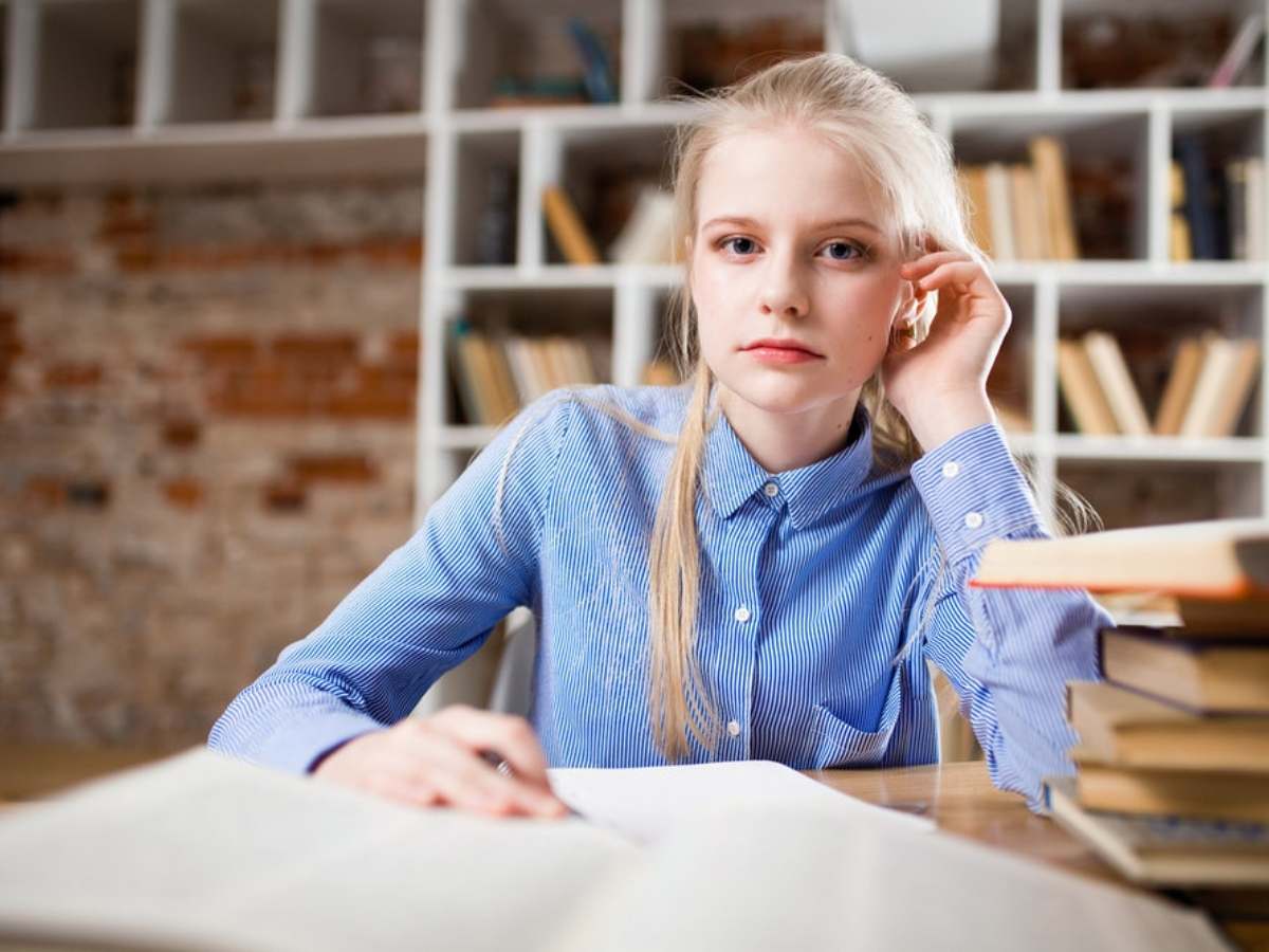 Girl with long blonde hair in blue button down shirt sitting at desk stacked with books and looking at camera for article Central Houston School Districts