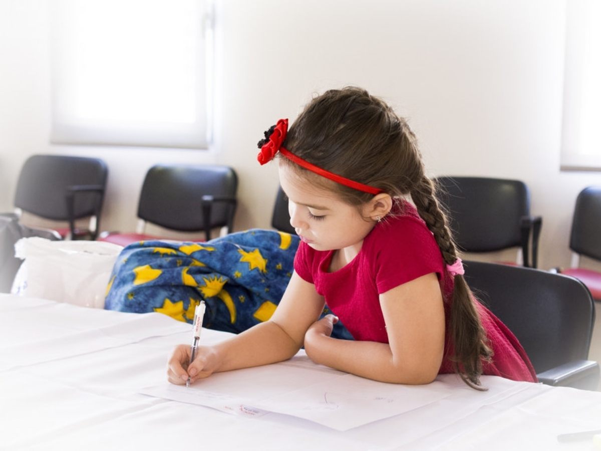 Young girl is bright pink dress with bright pink bow in hair and hair in braid sitting at a table writing for article Primrose Schools at Greenway Plaza & Upper Kirby