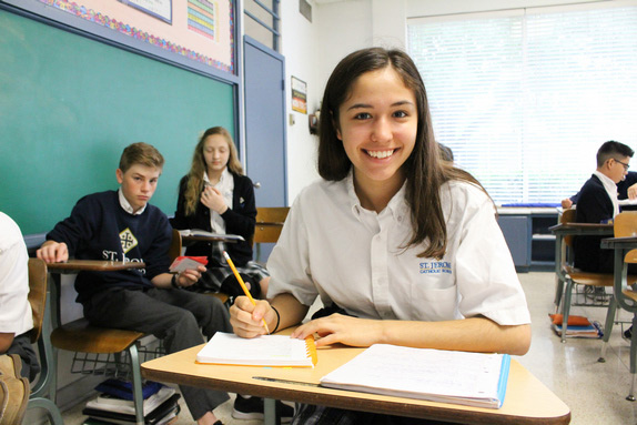 St. Jerome Catholic School - 1 girls sitting at desk in classroom in foreground smiling - other kids at desks in background for article St Jerome Catholic School for newcomers moving to Houston.