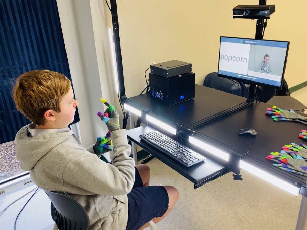 Boy sitting in front of AI camera/computer system with special gloves on to help read his hands as he practices sign language for article Fort Bend Christian Academy ASL Program