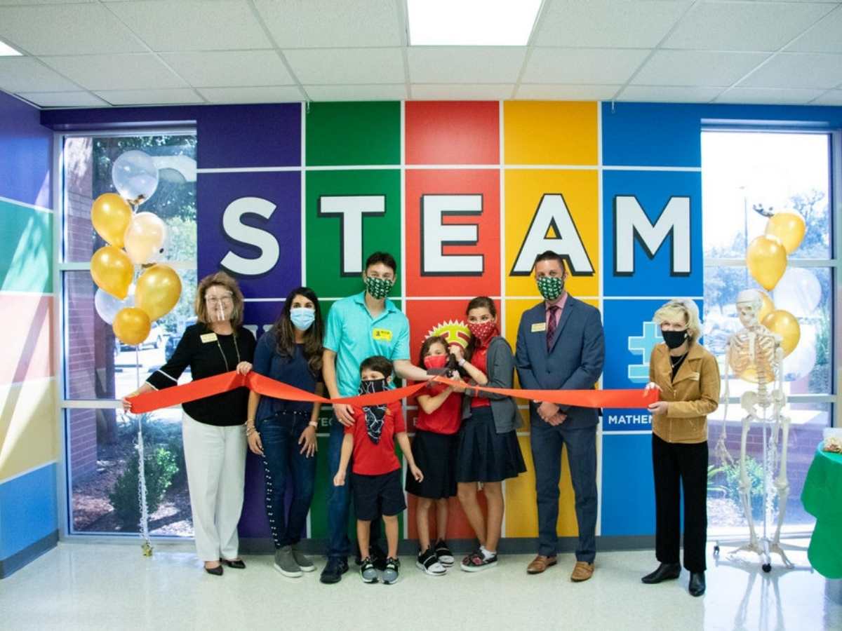Group of people standing with red ribbon in front of them and STEAM painted in color on wall behind them for article Fort Bend Christian Academy Family Celebrates Polasek STEAM Lab