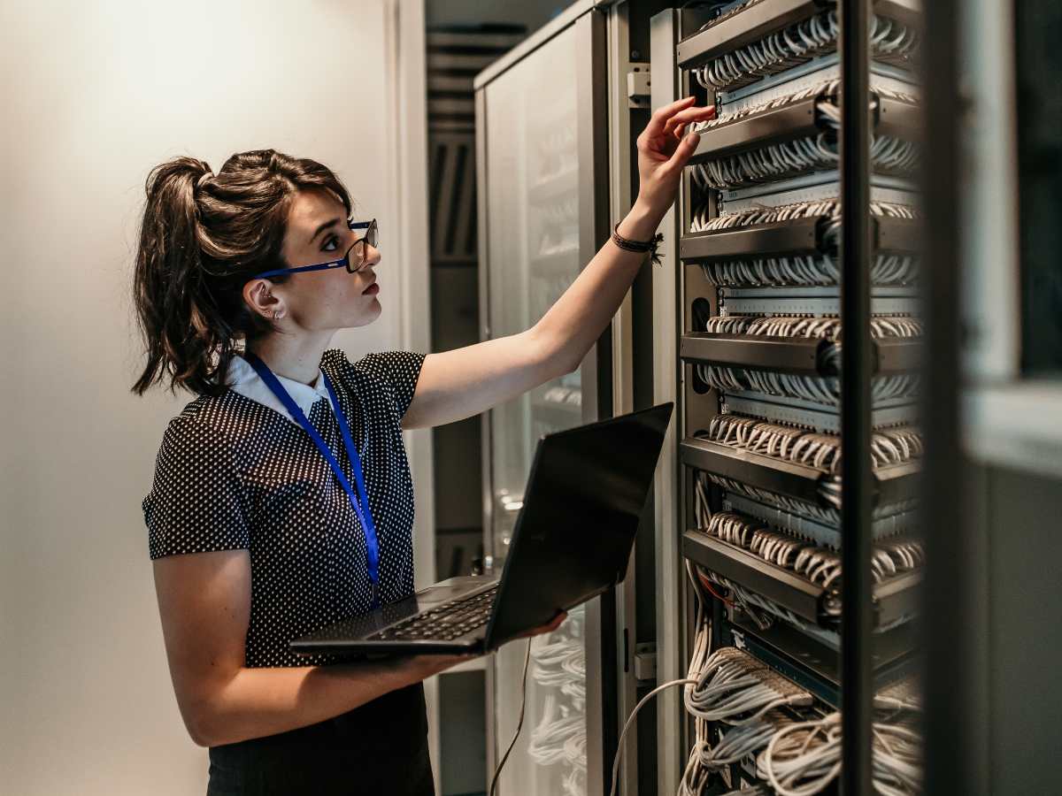 Woman with glasses standing holding an open laptop and working on computer network in server room for article Houston Women in Tech: Ranked in Top 3 for newcomer moving to Houston.