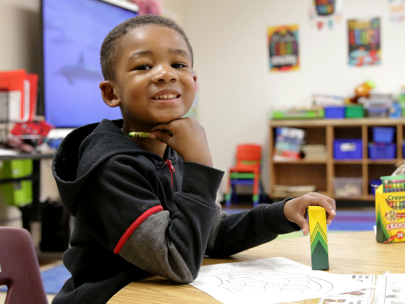 young male student smiling at the camera holding a crayon while working in class