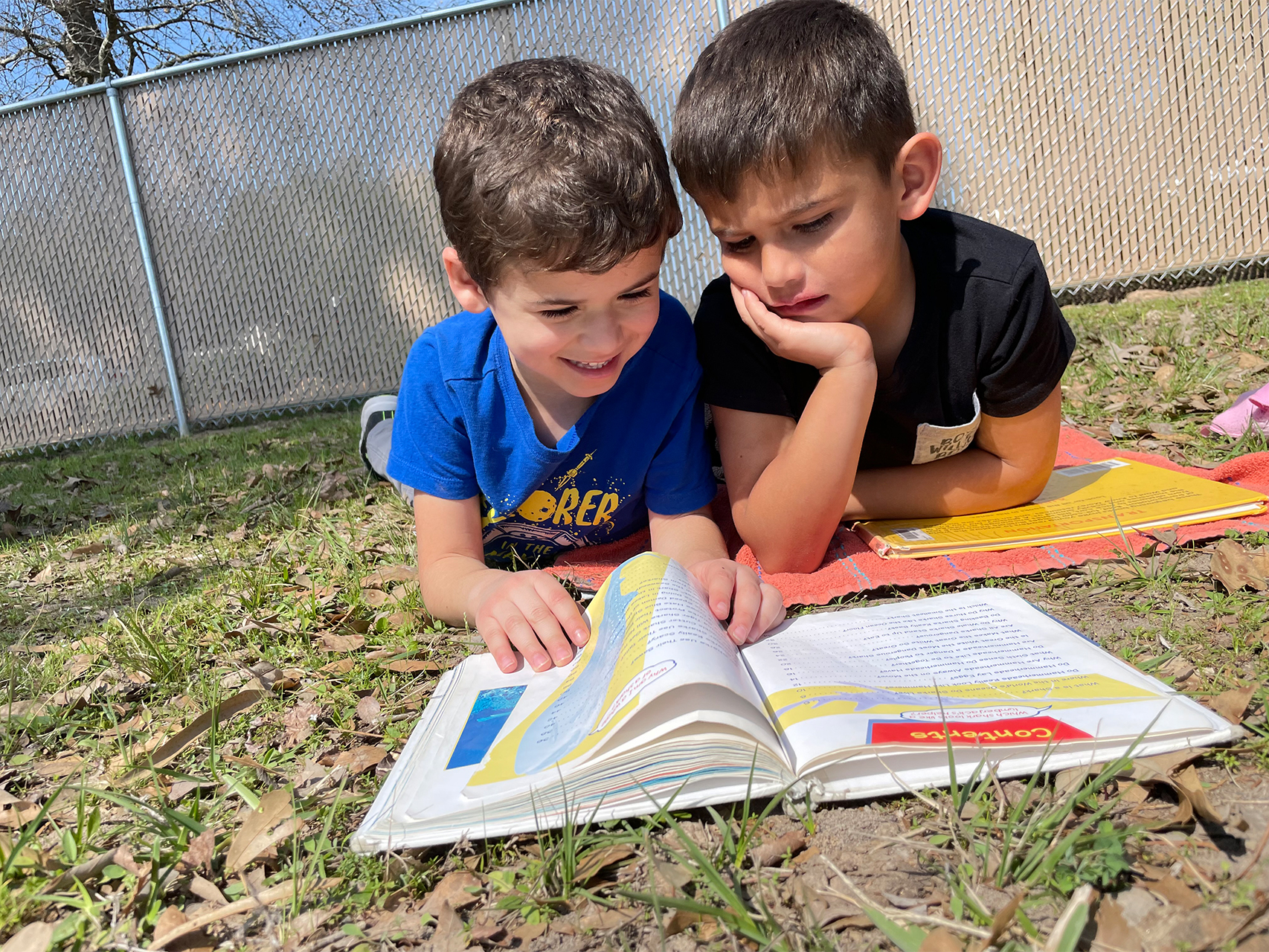 two young male students laying on the ground reading a book together and laughing