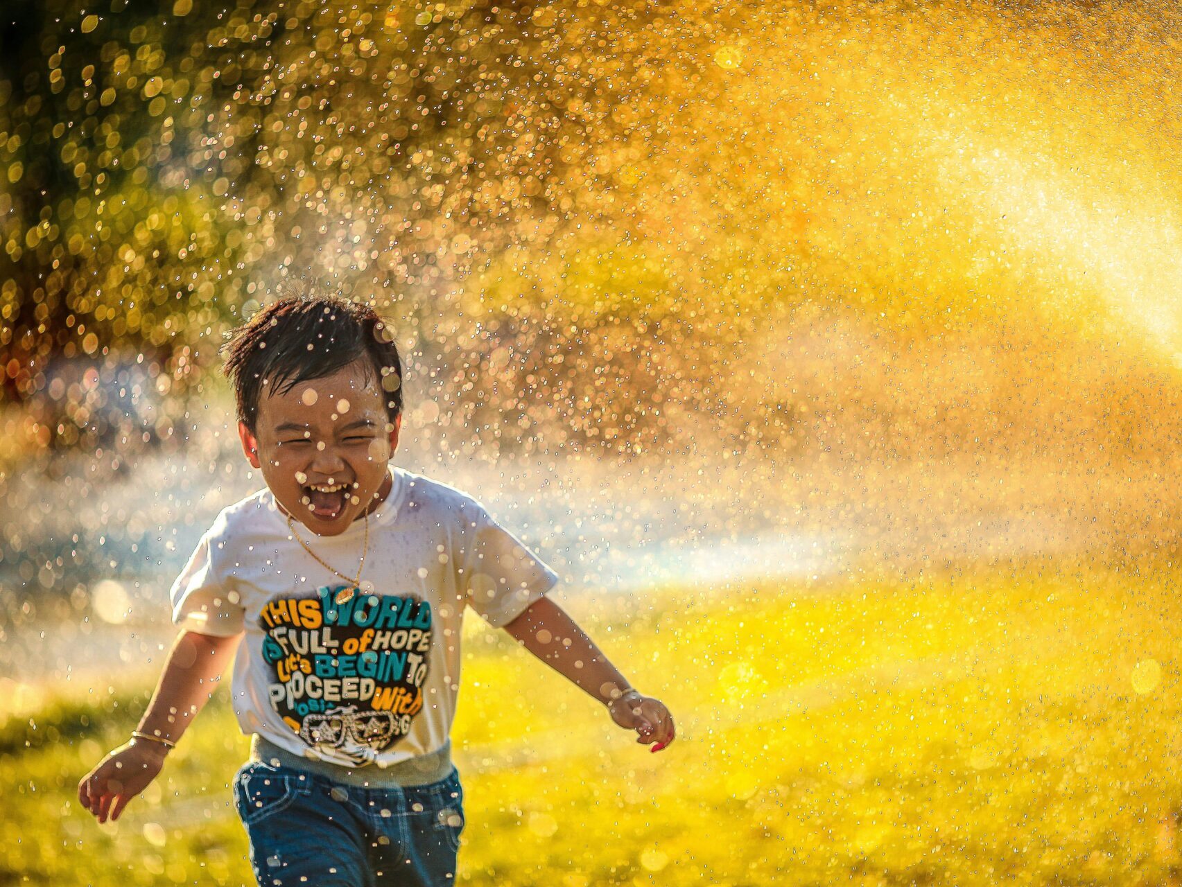 boy running through sprinkler in yard on grass, smiling