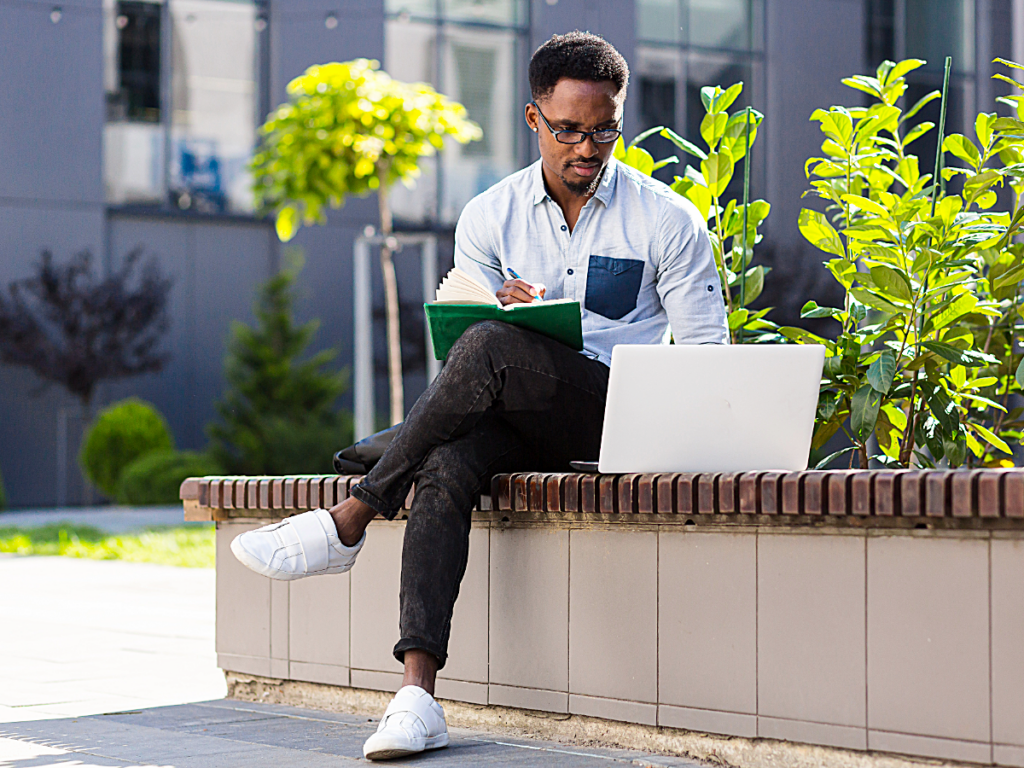 Male sitting outside working on computer and writing in his notebook.