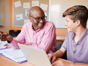 Man sitting at desk tutoring a male student sitting next to him with his laptop open on the desk.