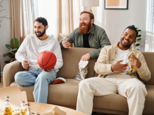 3 males - 2 sitting on couch, 1 behind the couch, drinking beer and watching a game together.