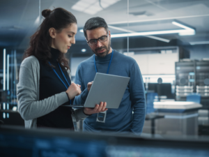 Male and female coworkers standing up looking over a laptop in a technology office for article 10 Leading Technology Companies in Houston for newcomers moving to Houston.