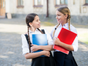 2 High school girls in private school uniforms holding books and looking at each other with school in background for article Top 4 Private High Schools in Houston for newcomers moving to Houston.