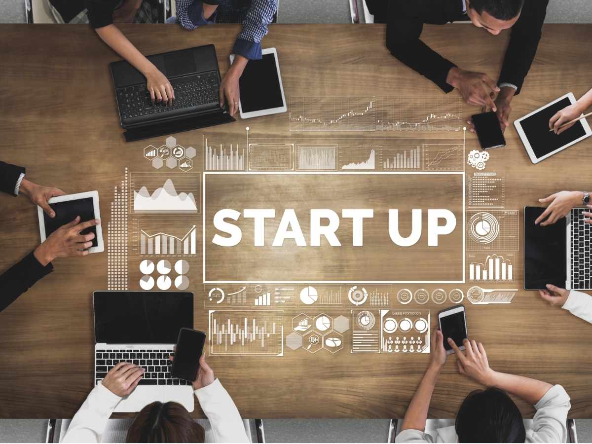 Aerial view of people sitting around wood conference table with their phones, tables, laptops - center of table has white text overlay that says start up with generic charts and graphs around the lettering for article Start a Business in Houston: 7 Smart Steps for Newcomers in 2025 for newcomers moving to Houston.