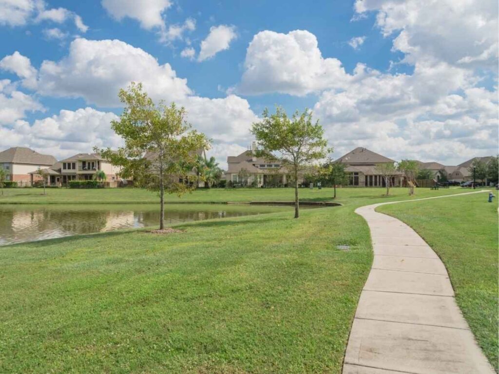 Suburban neighborhood with walking path around lake with green grass and large homes in background for article Living in Pearland, TX in 2025 for newcomers moving to Pearland, TX and Houston area.